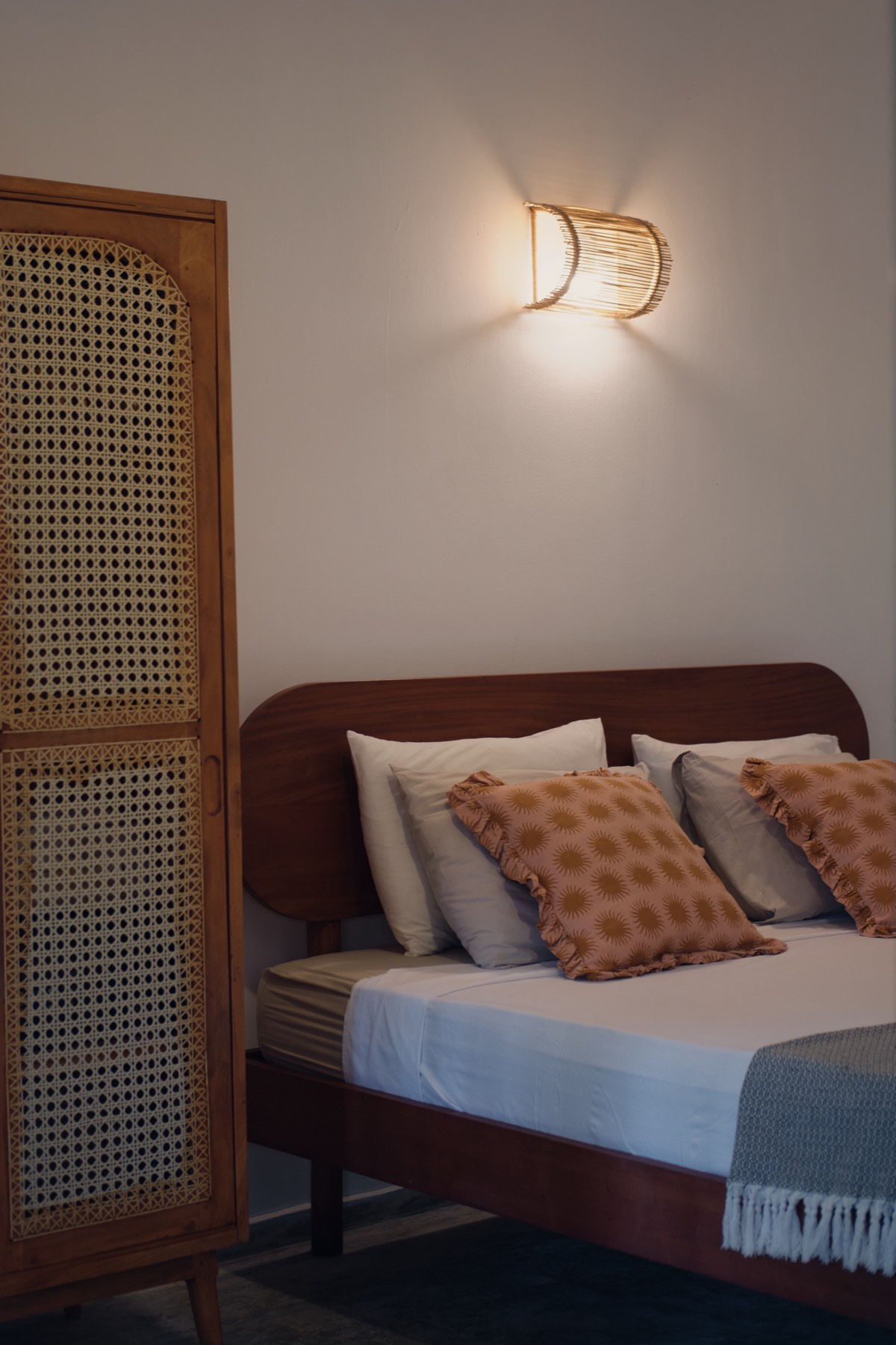 A room by lamplight — a rattan wall light over the headboard, pillows on the bed, the cane wardrobe beside it.