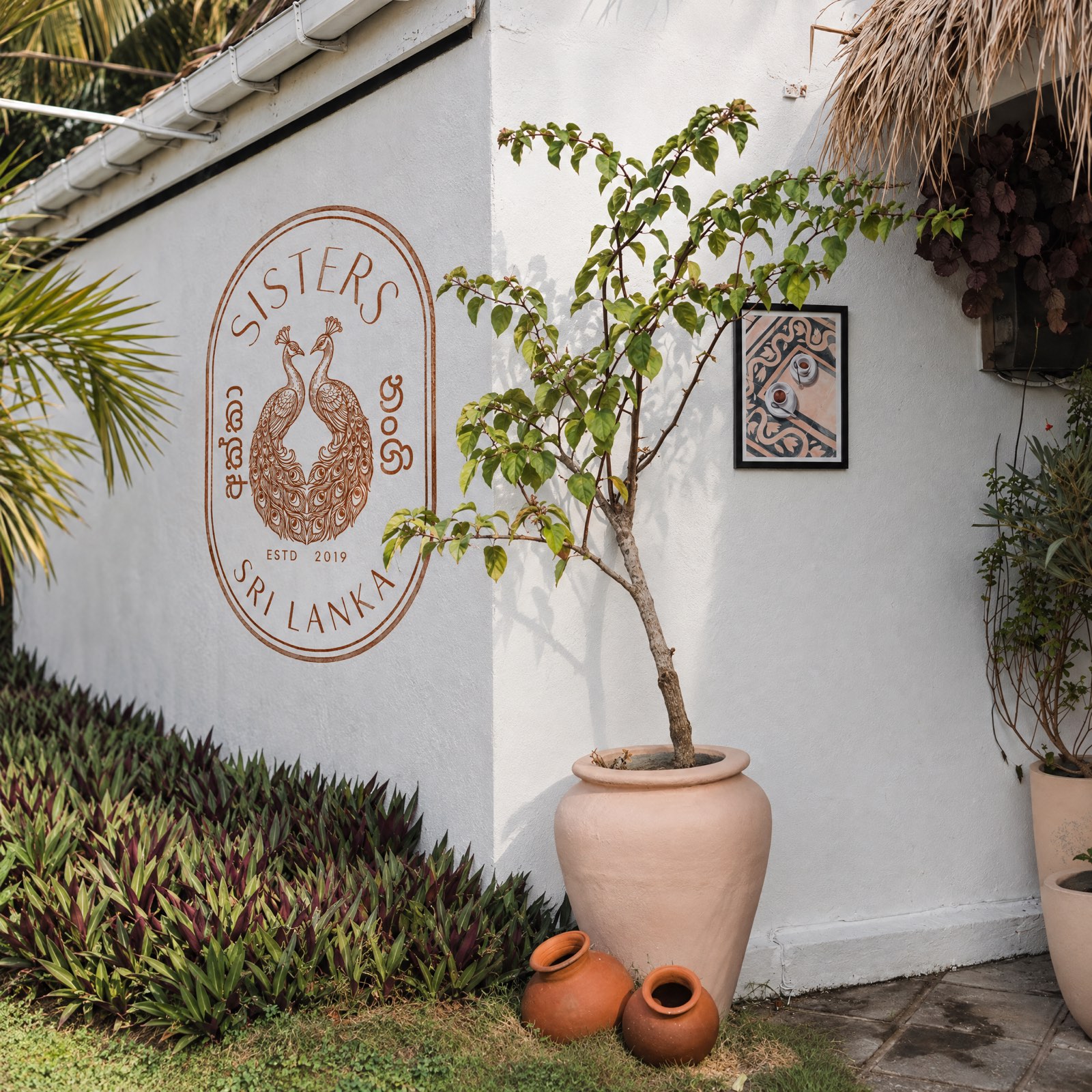 The Sisters crest hand-painted large in terracotta on a white exterior wall, beside a framed print and a potted tree.