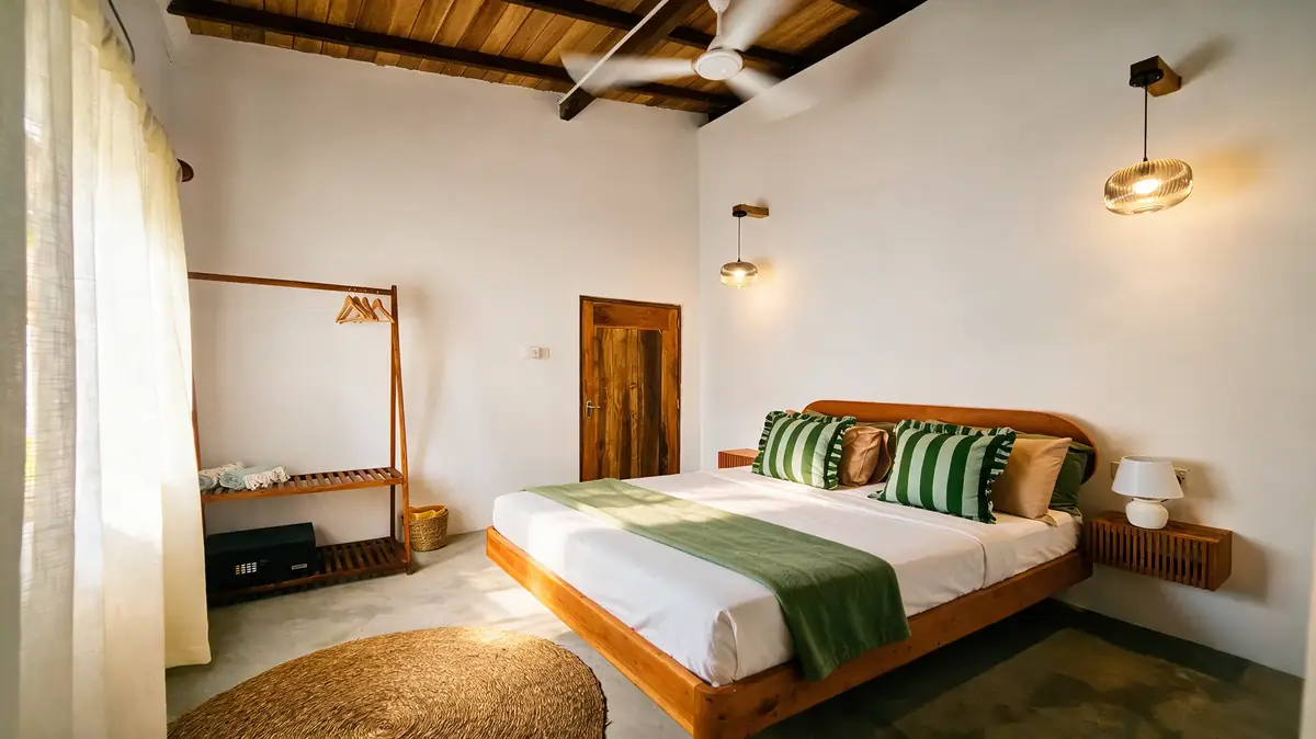 A guest room at Sisters Away — teak bed, green striped bolsters, a timber ceiling, a clothes rail against a white wall.