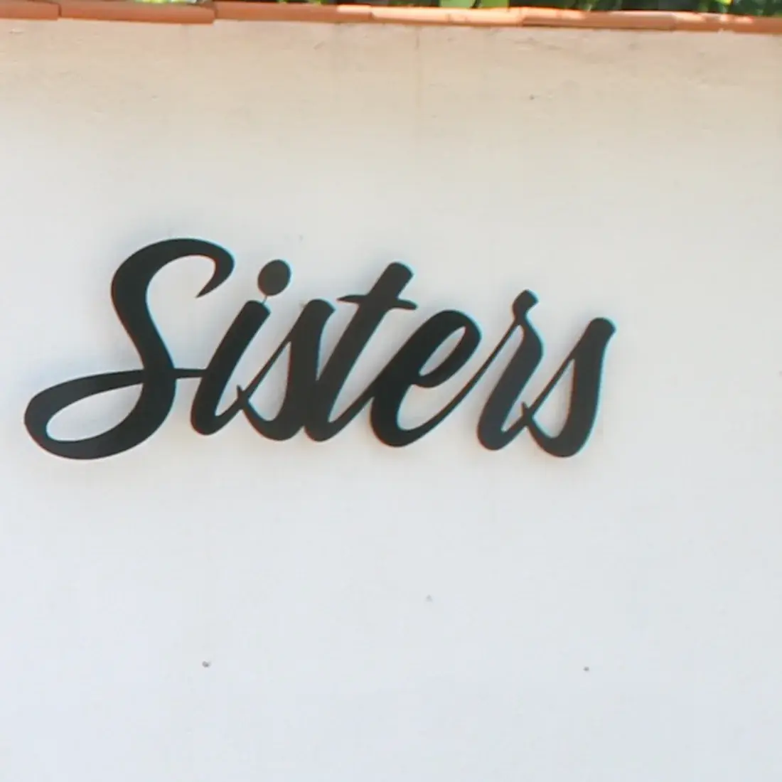 The old identity — a flowing script wordmark reading Sisters, cut in black and fixed to the white gate wall.