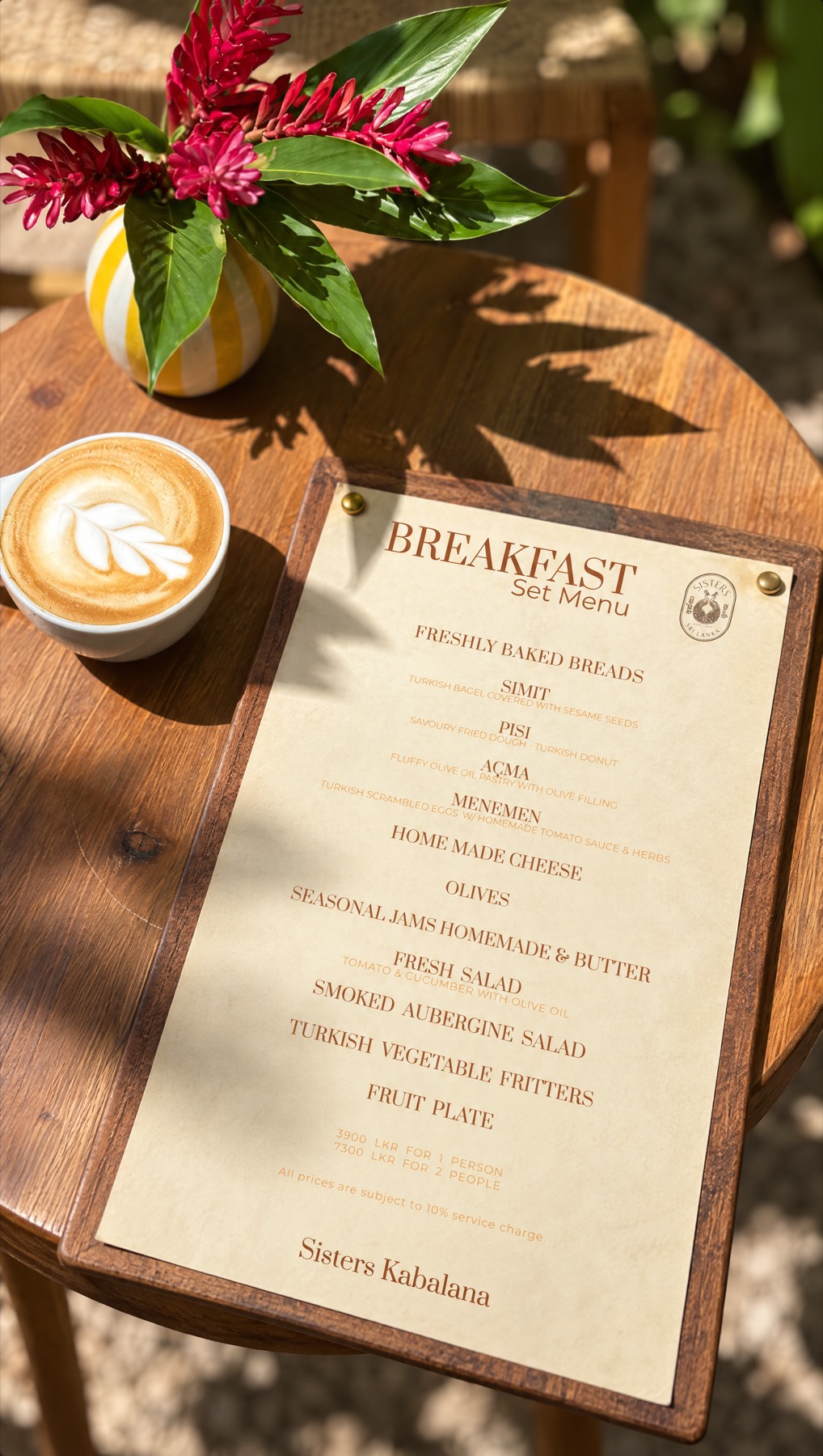 The same set menu clipped to a wooden board on a café table, beside a cappuccino and a vase of red ginger flowers.