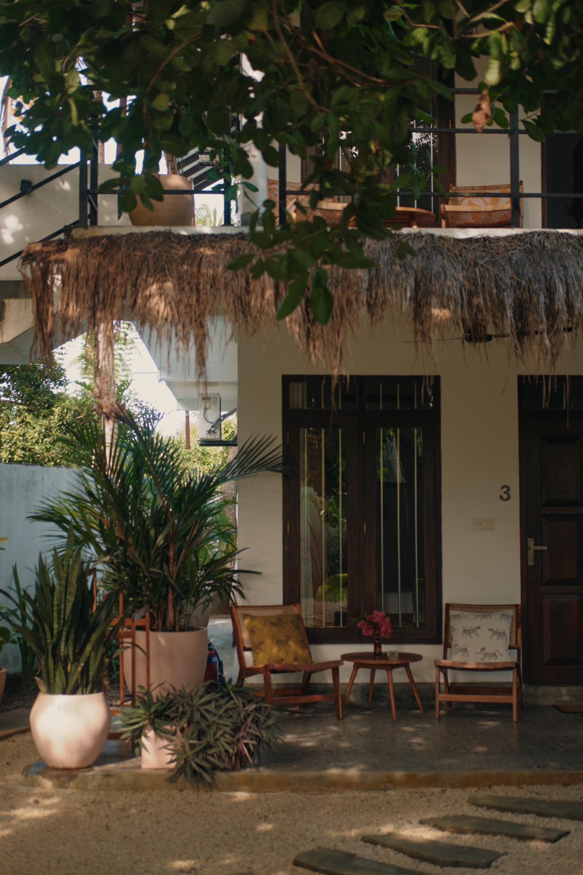 A guesthouse room at Sisters Away — dark timber door and shutters, thatched eave, planter chairs on the veranda, palms crowding in.
